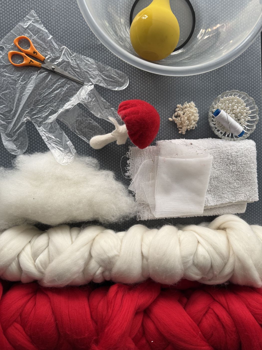 Red and white merino wool, a yellow water sprinkler, bowl and felting tools laid out